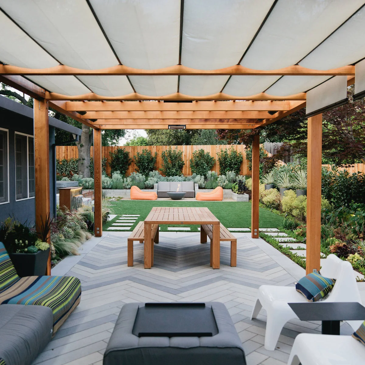 White fabric panels span a wood pergola above a picnic table and lounge seating on patterned paving.