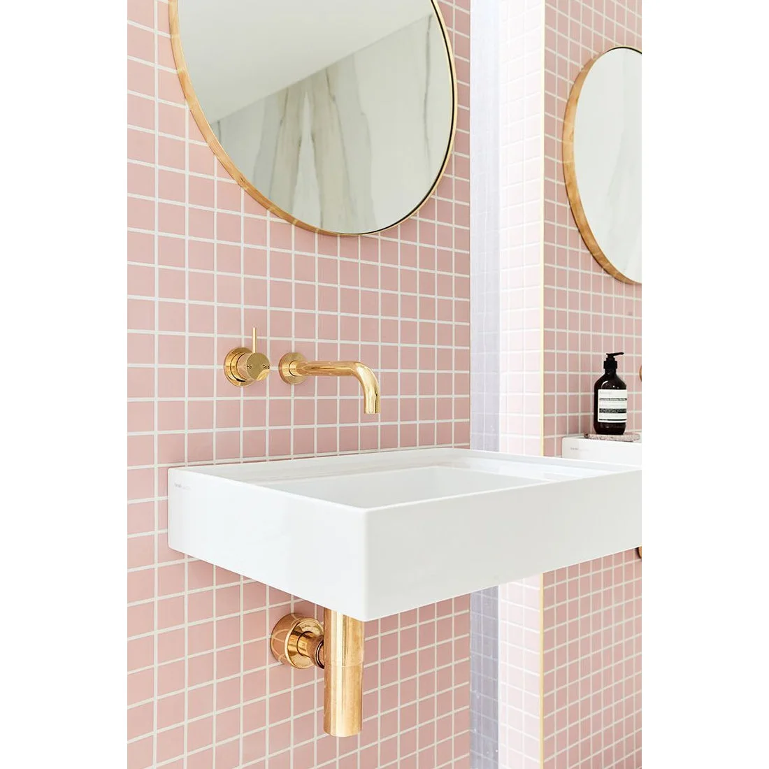 Small pale pink square tiles surround a wall-mounted white sink beneath two round gold-framed mirrors