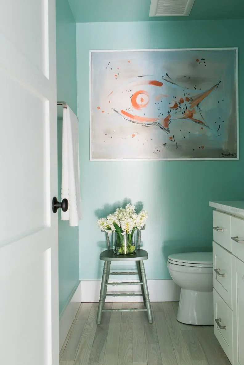 Mint walls surround a compact white vanity and toilet, with a large framed abstract picture, pale floor and small green stool holding flowers.