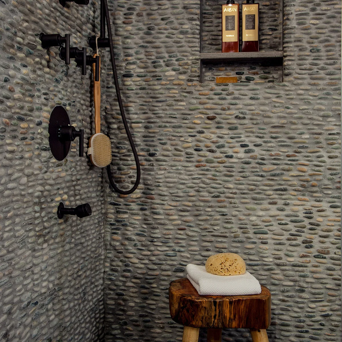 A close view of pebble-clad shower walls with dark fittings, a recessed shelf and a small wood stool holding folded towels.