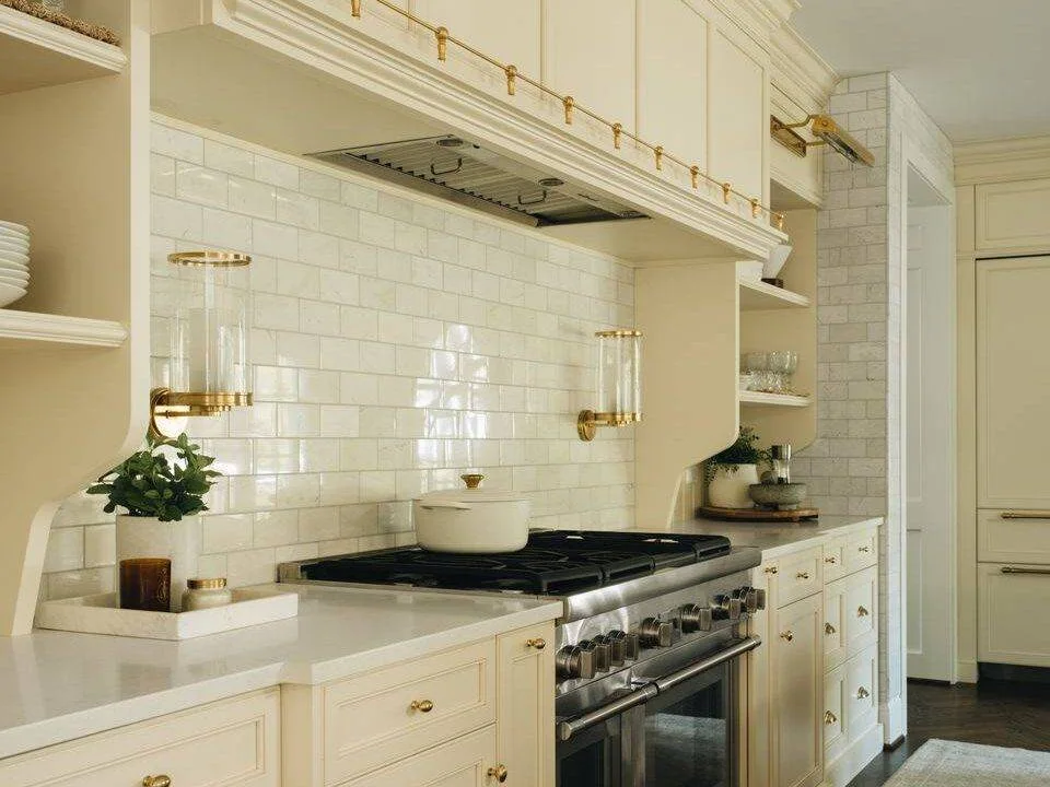 Pale marble subway tiles sit behind a range beneath a cream hood with warm-metal details and open side shelves.