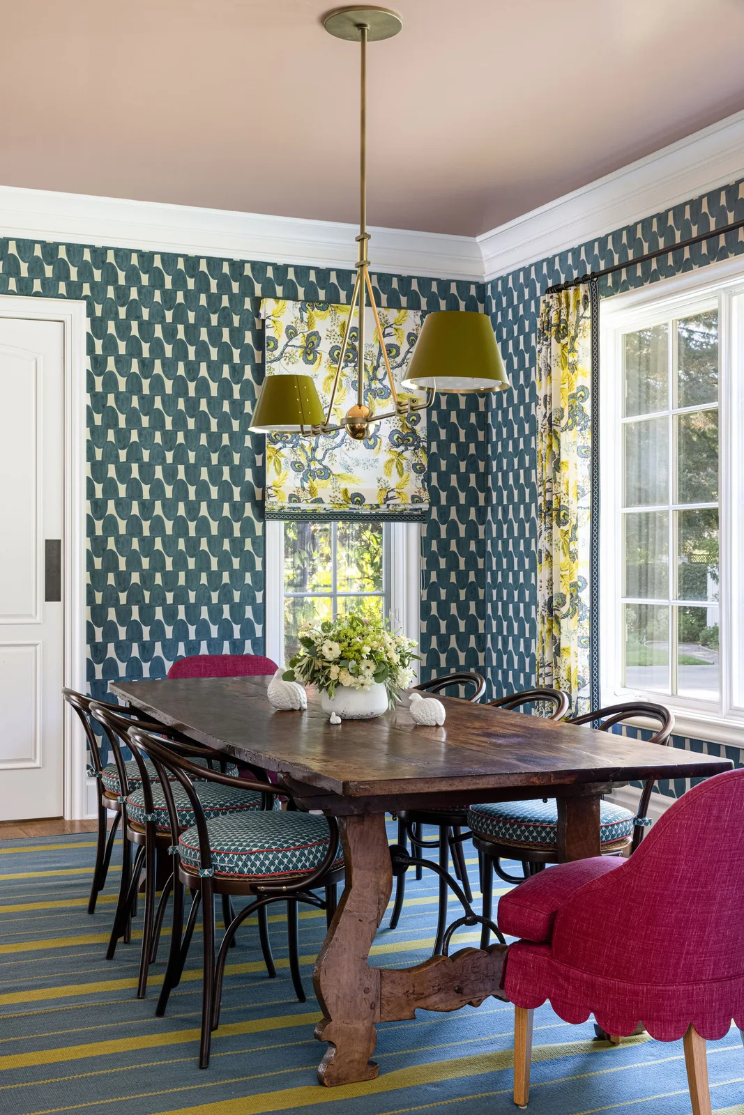 Blue geometric wallpaper surrounds yellow-and-blue patterned blinds above a striped rug, dark bentwood chairs and pink end chairs
