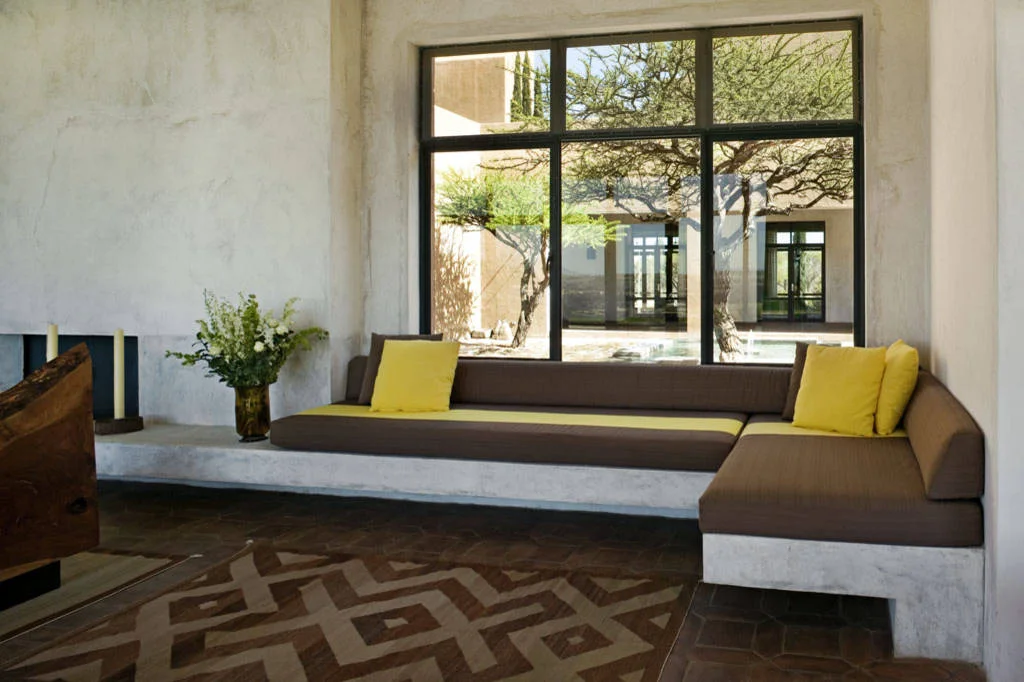 L-shaped built-in bench with brown seat cushions and yellow pillows below a large dark-framed window.