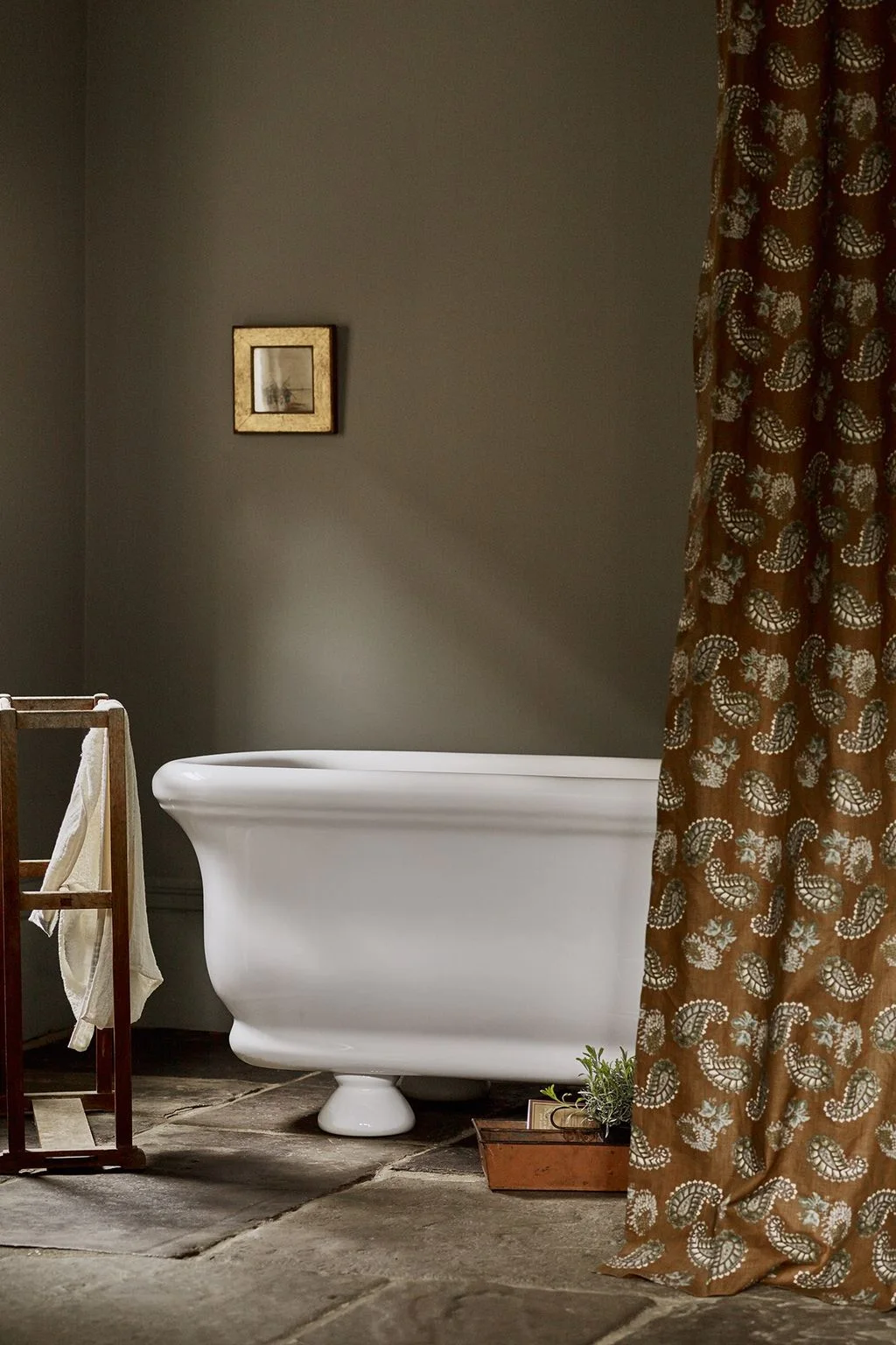 Muted olive walls sit behind a white freestanding bath, a copper-toned patterned curtain and a wood towel ladder on a textured stone floor.