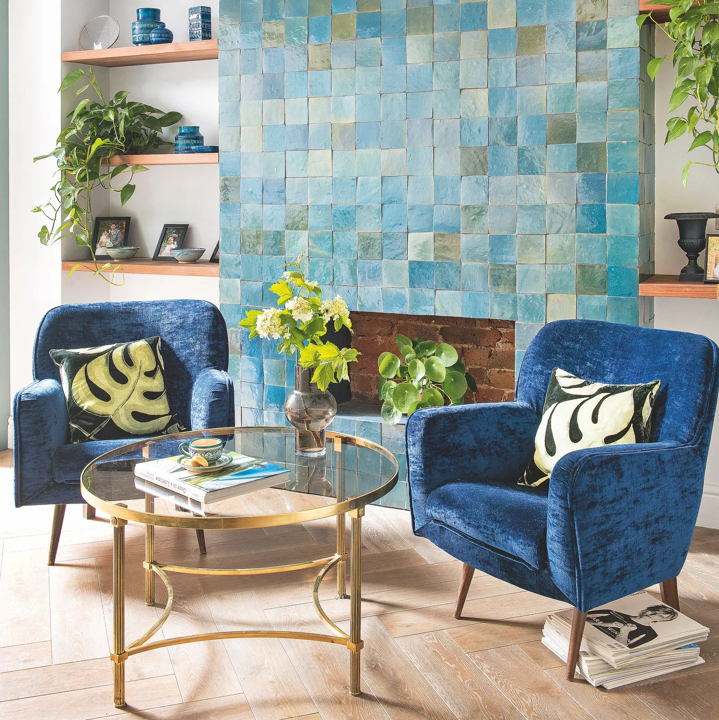 Blue, turquoise and muted green square tiles cover a chimney breast beside wood shelves and blue armchairs, with plants in the unused opening.