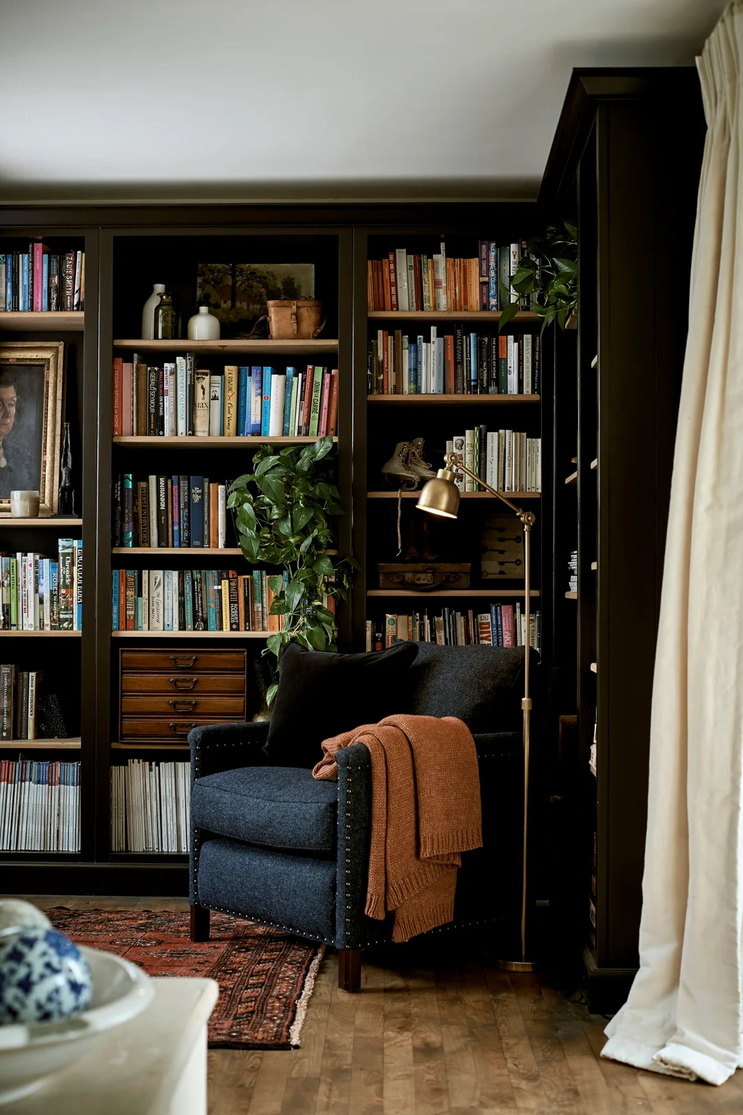 A dark upholstered chair with an orange throw sits before dark bookshelves with wooden shelf edges beside a gold-toned adjustable lamp