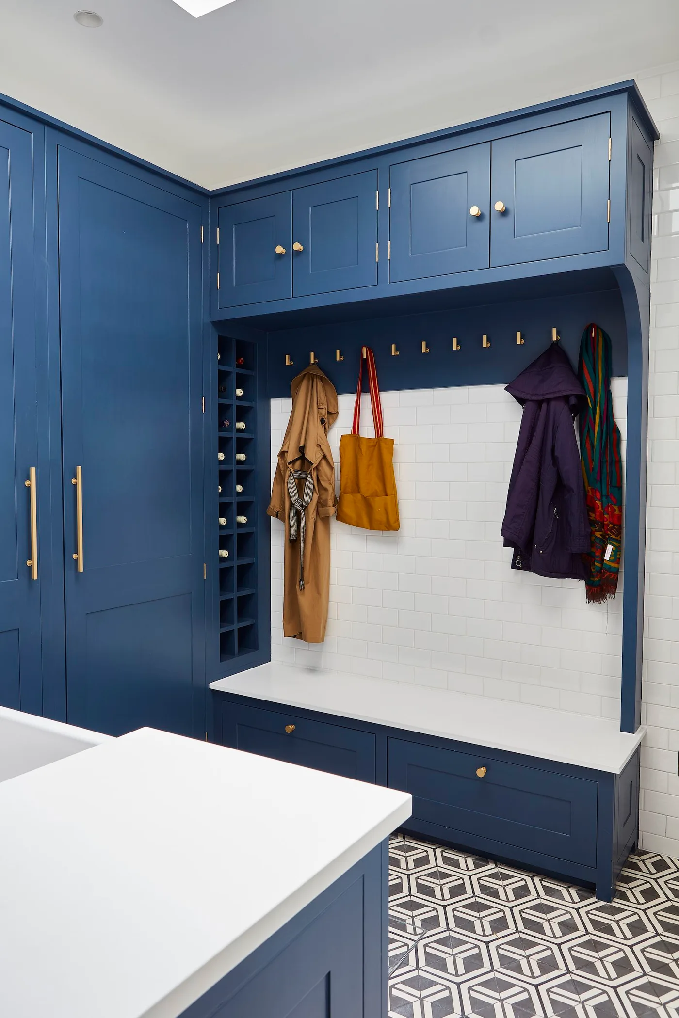 Blue corner mudroom cabinets with tall cupboard, upper cupboards, pale bench, coat hooks and geometric floor.