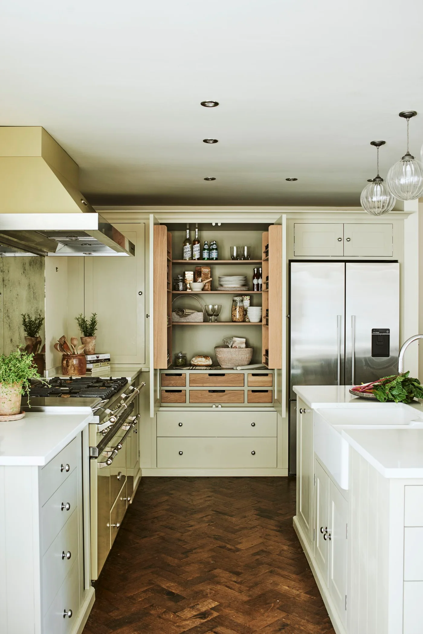 Cream framed kitchen cabinetry around an open wooden pantry beside a stainless range, refrigerator and dark herringbone floor.