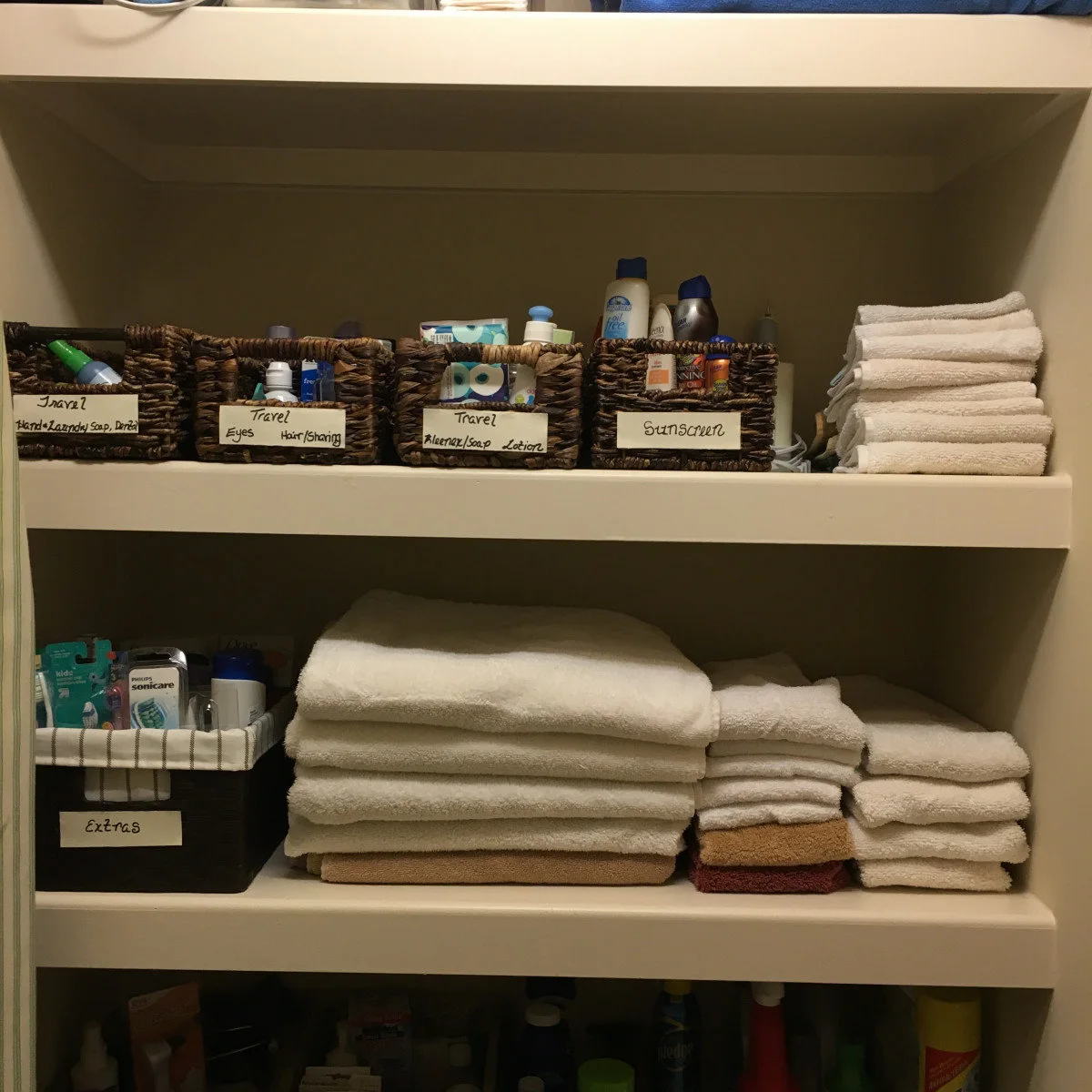 Open closet shelf with labeled travel toiletry baskets above stacks of white and beige towels