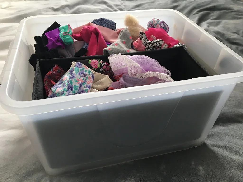 Clear storage tub with black rectangular inserts separating colorful doll clothes.