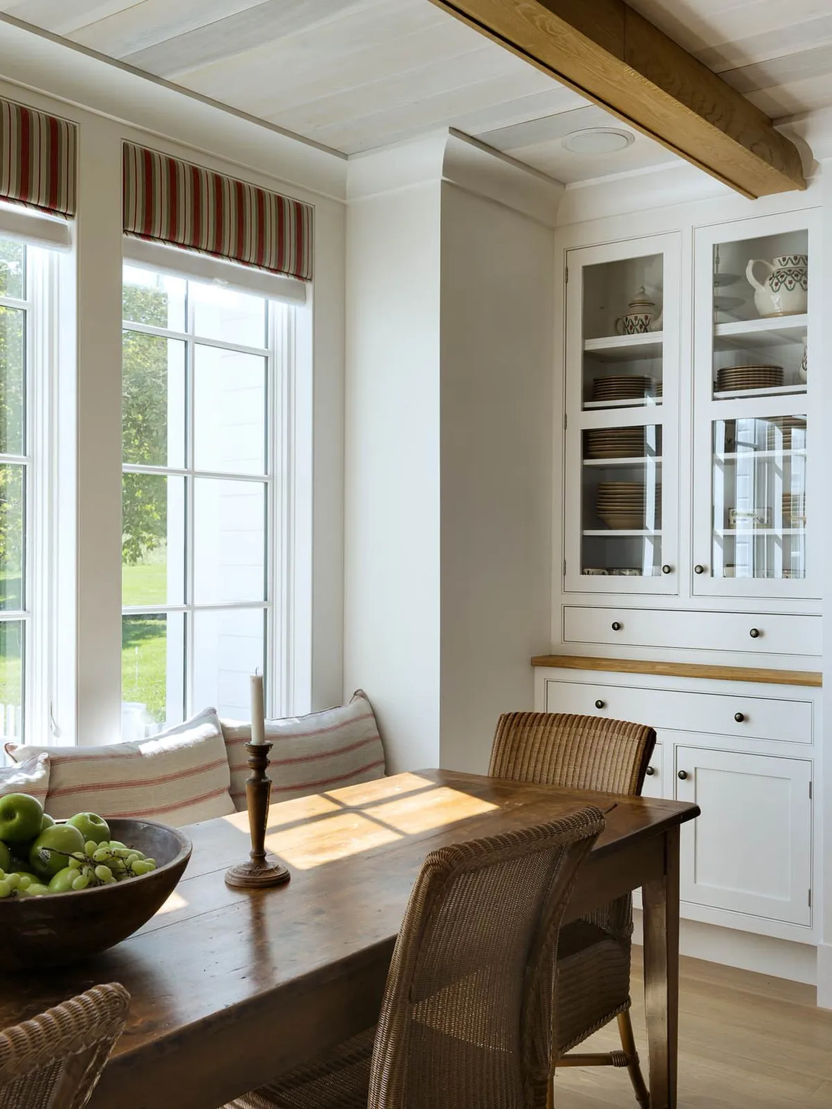 A timber dining table sits near pale glass-front cupboards and a bright window.