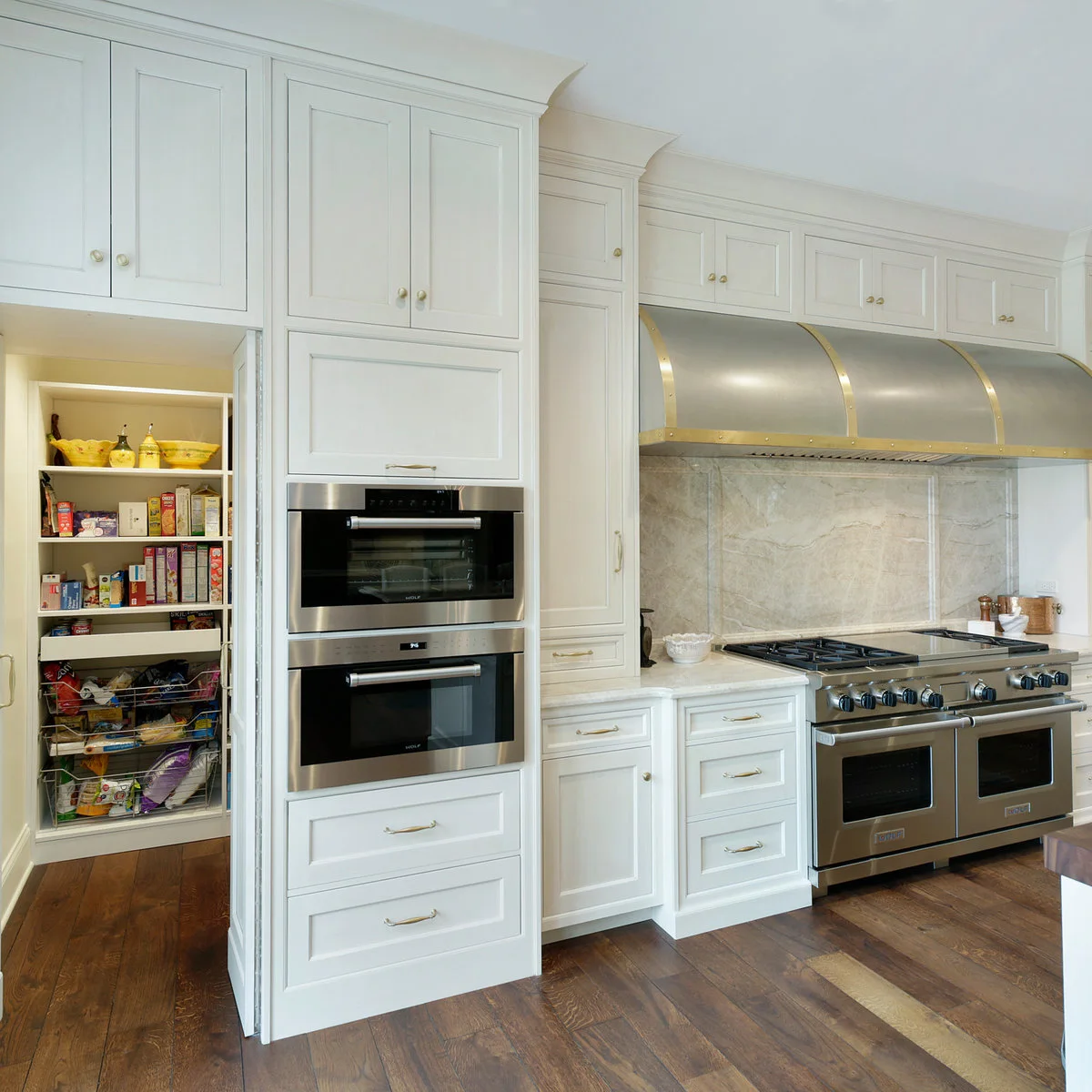A tall white door opens beside stacked ovens to reveal pantry shelves behind a continuous white kitchen cabinet run.