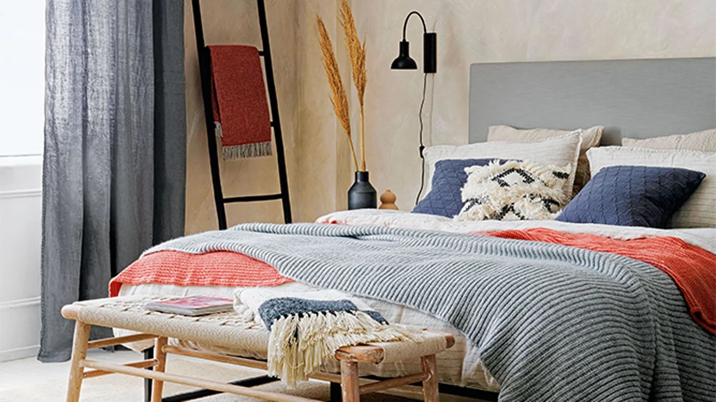 A gray headboard sits behind blue cushions and pale bedding with coral and gray throws beside a dark towel ladder and wooden bench