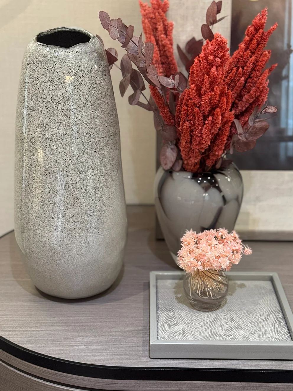 A tiny pale pink dried flower bunch sits in a small glass vase on a tray beside taller gray and patterned vessels with red-toned stems.