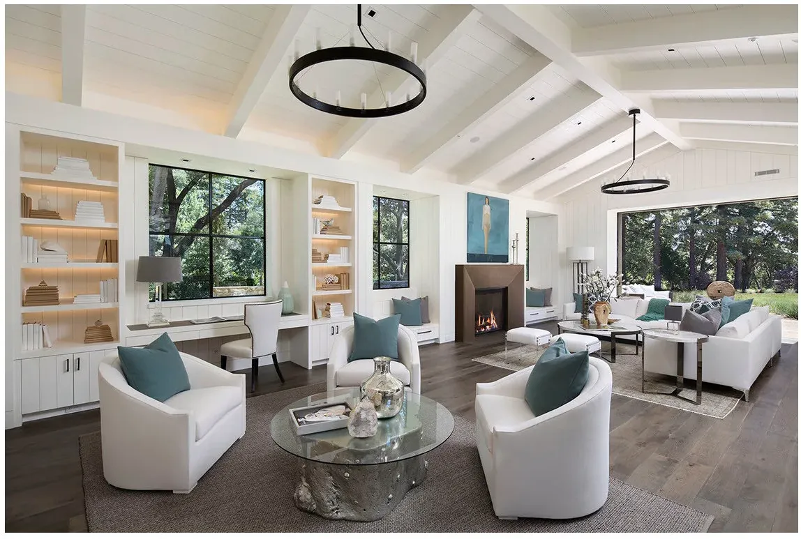 A built-in desk beneath a window between illuminated bookcases in a large pale living room with several seating groups.