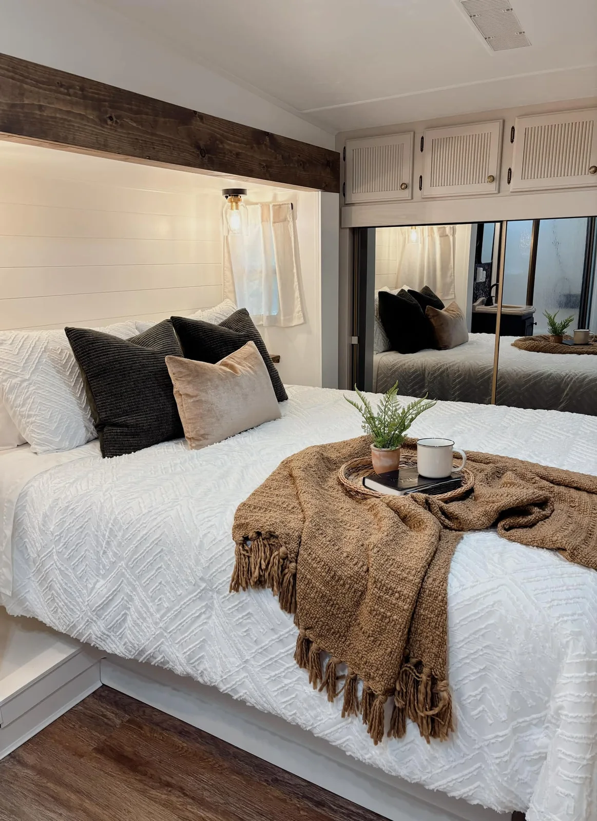 RV bedroom with white bedding, dark pillows, two wall lights and mirrored wardrobe doors.