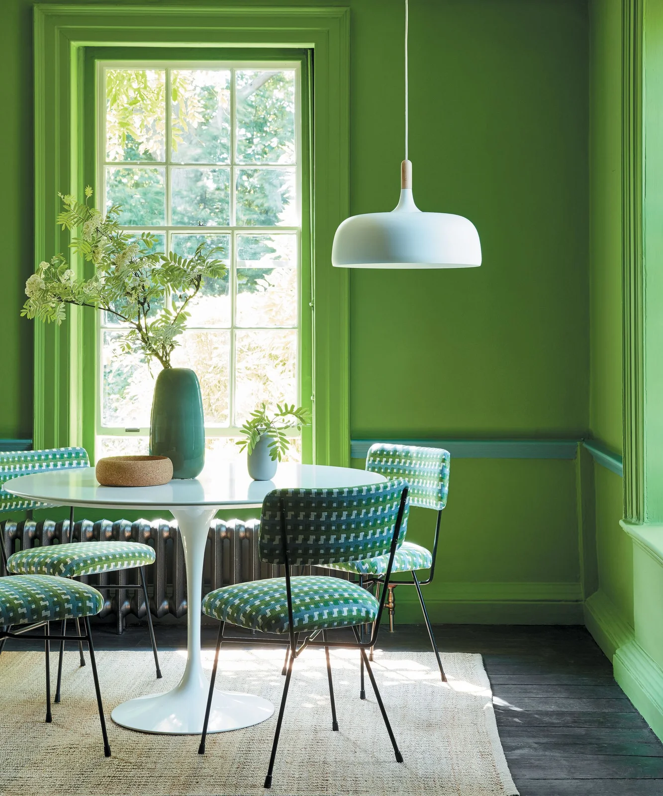 Bright green walls and trim surround a pale round table with patterned green chairs beneath a pale pendant beside a tall window