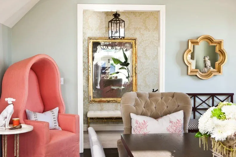 Tall coral hooded chair beside a dark dining table and a pale tufted chair, with framed mirrors and a lantern light beyond.