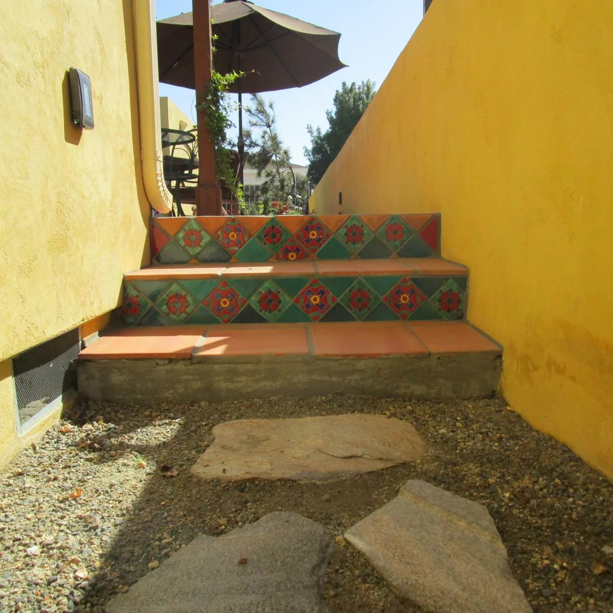 Short outdoor steps with green and multicolored patterned riser tiles between yellow walls beside a tiled patio.