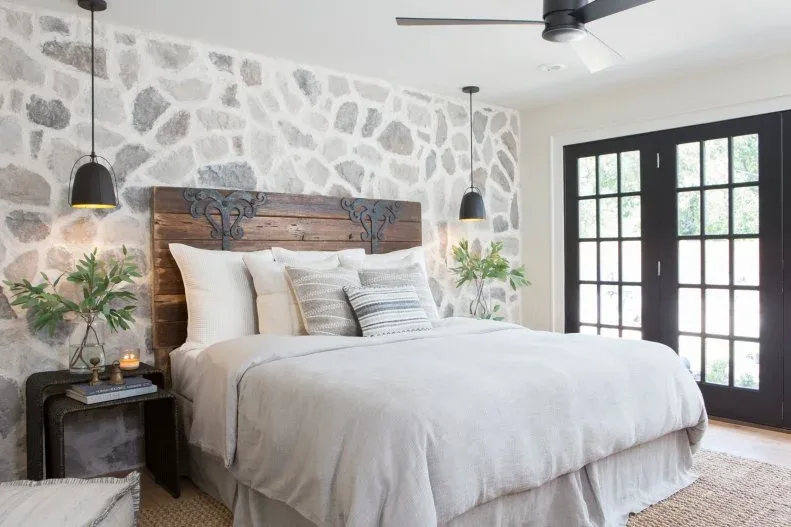 A timber headboard with dark decorative brackets stands against an irregular stone wall beside pale bedding and black doors.