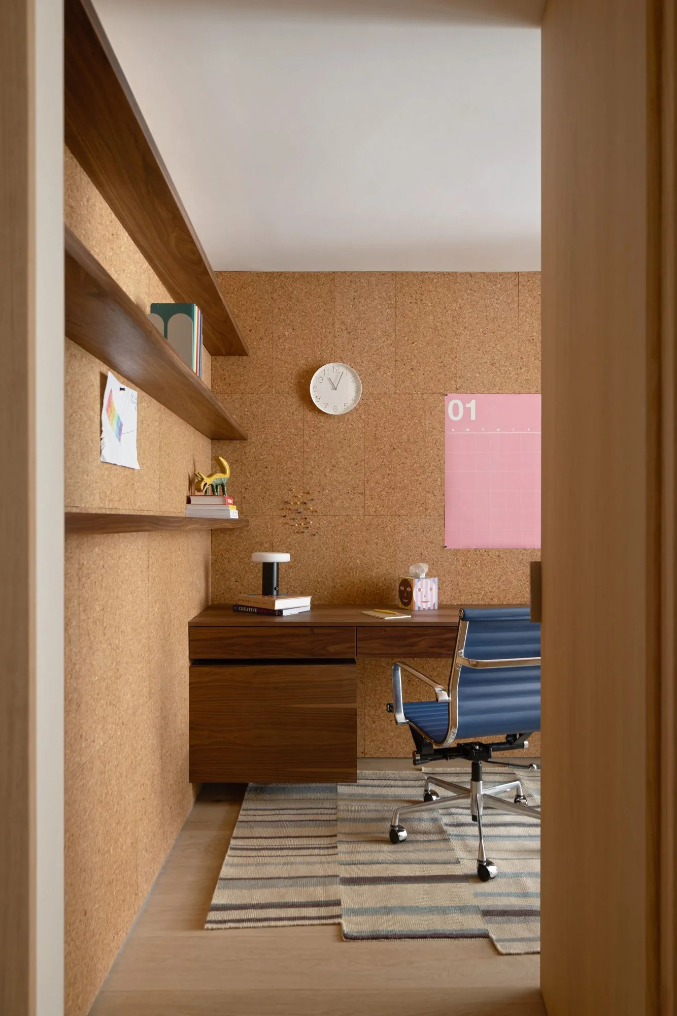 Home office with textured tan walls, wooden desk and open shelves, blue rolling chair, striped rug, round white clock and pink wall calendar.