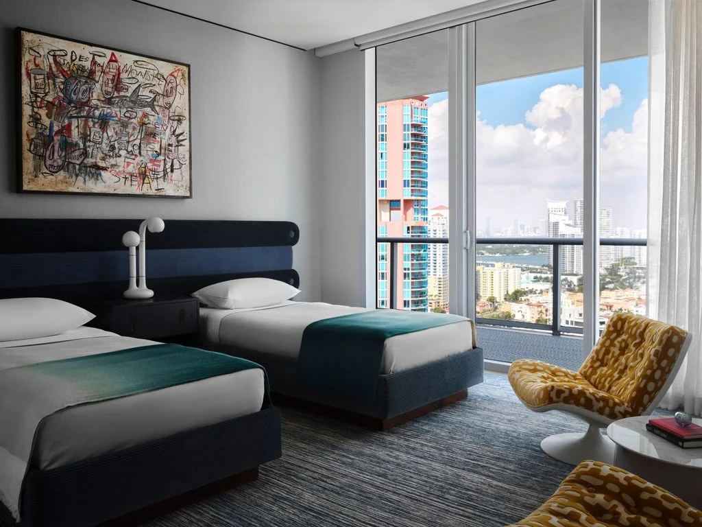 Two single beds with dark headboards and green throws stand below one abstract picture beside broad windows and two patterned lounge chairs