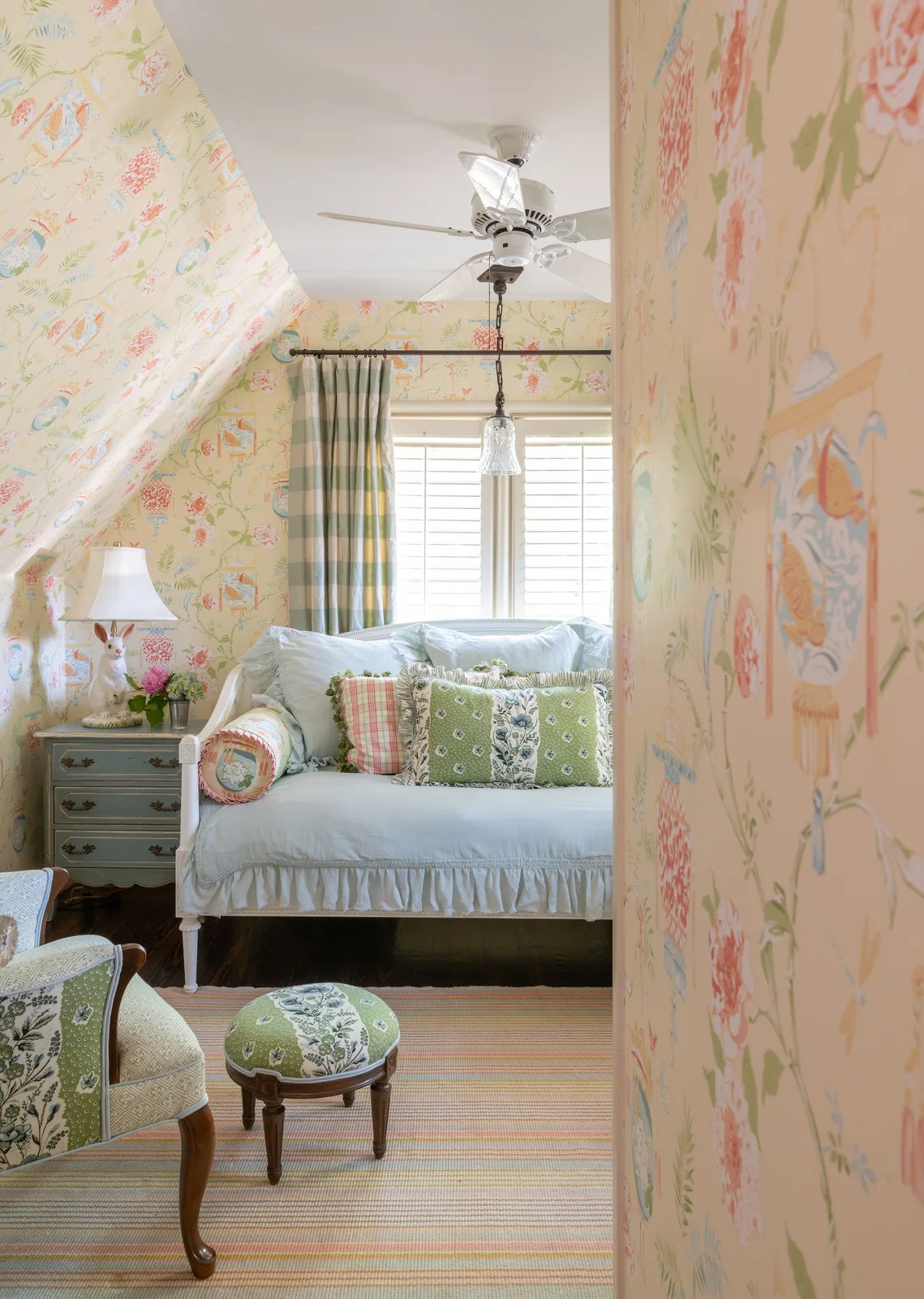 Pale yellow patterned wallpaper lines a sloping room around a white daybed with a blue cover, pastel cushions and striped curtains.