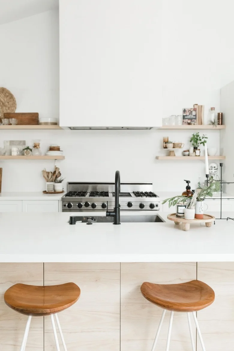 A white kitchen has pale wood island fronts, wood-seat stools, a black faucet, narrow open shelves and a broad plain hood above the range.