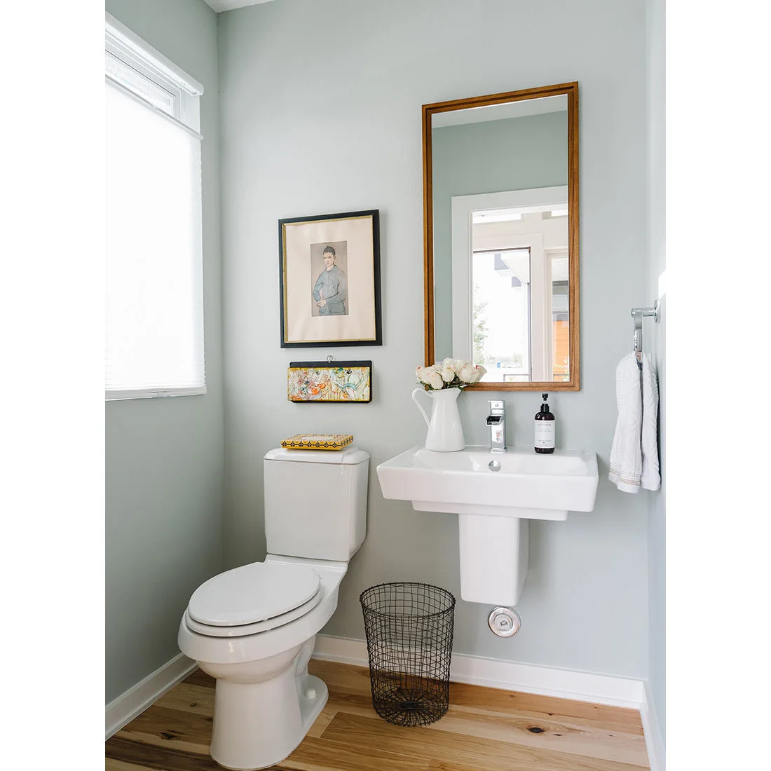 Pale green walls frame a white wall-mounted basin and toilet, with a wood-edged rectangular mirror, two framed pictures and a pale wood floor.