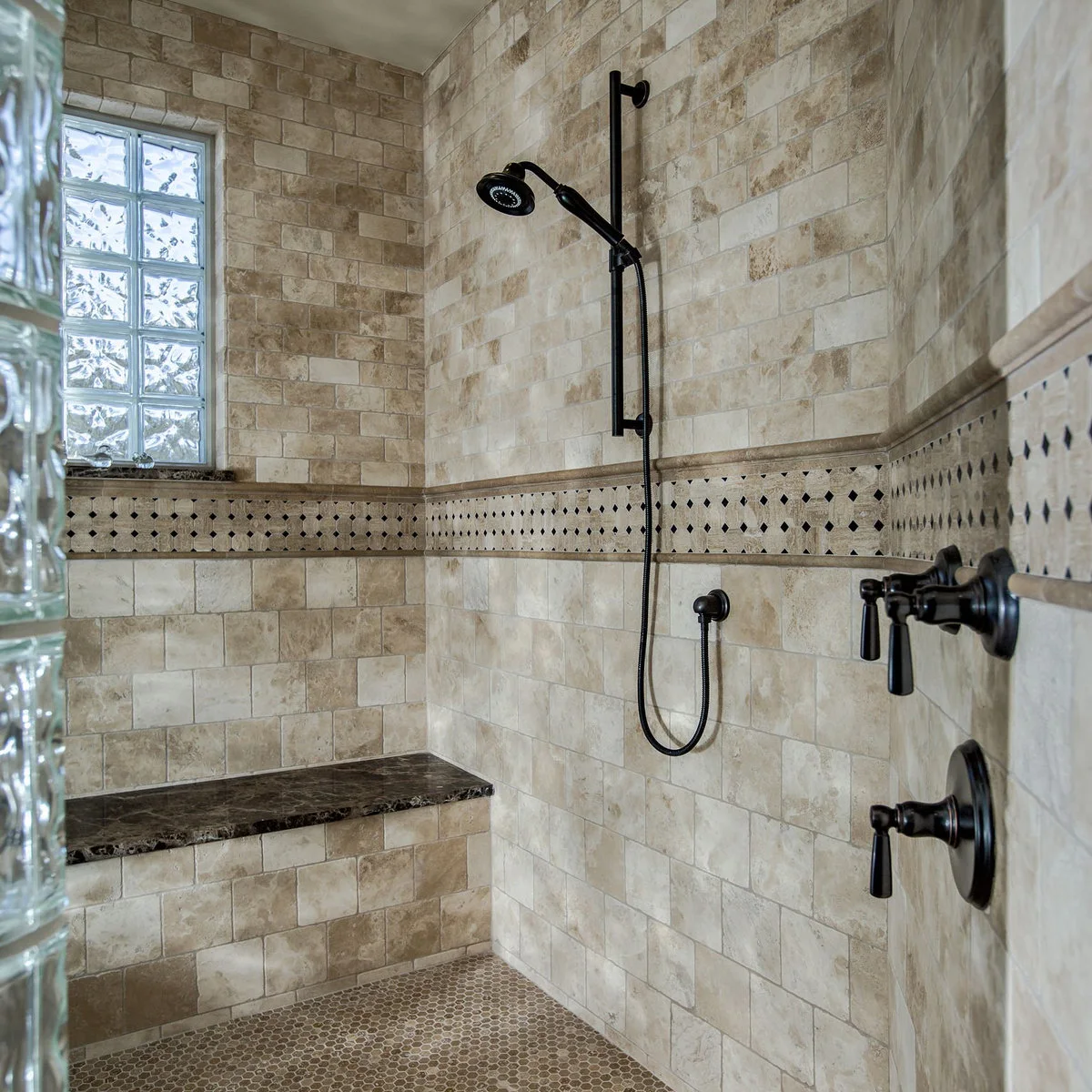 A pale tiled shower with a dark hand shower and controls, a long bench, patterned border and glass-block window.