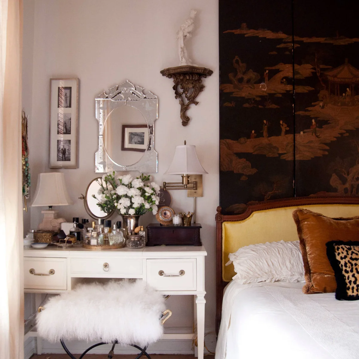 A pale dressing table with small bottles, two mirrors and a soft stool stands beside a yellow upholstered bed