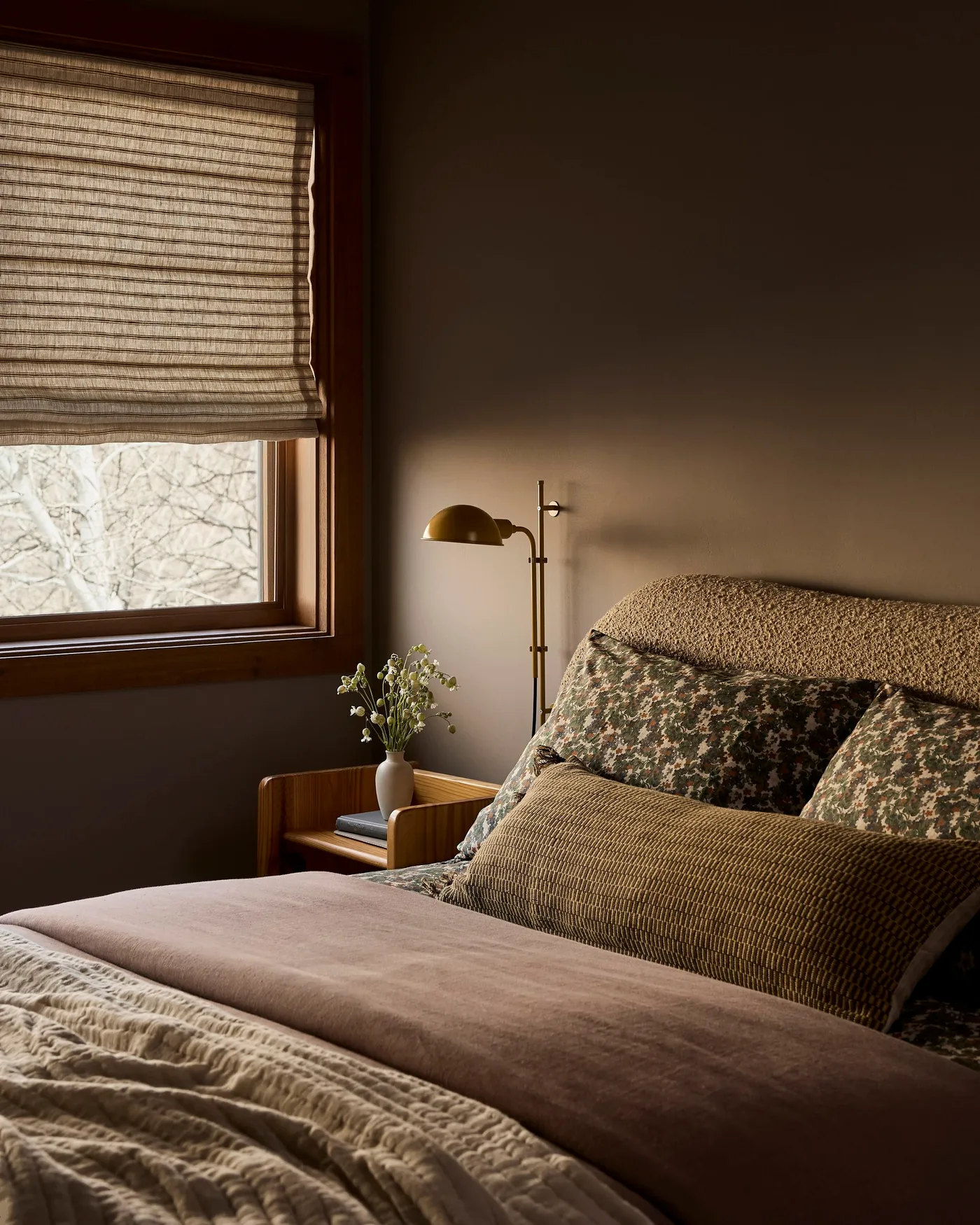 Brown bedroom with a textured taupe headboard, muted pink blanket, olive patterned pillows, striped cushion and woven window shade