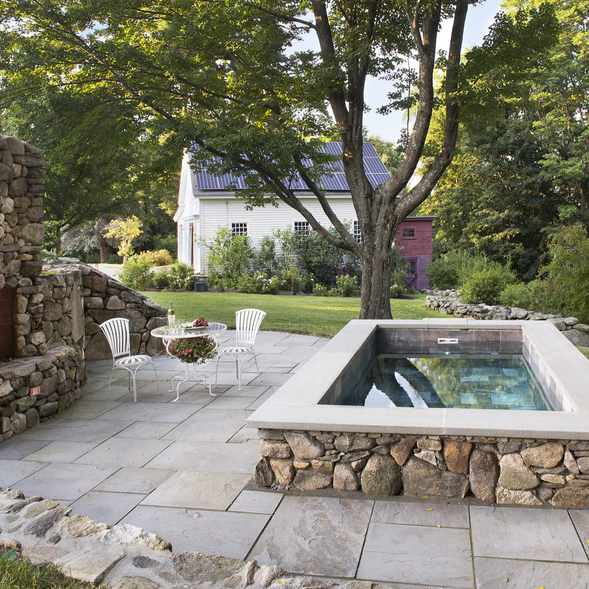 A raised rectangular plunge pool with an irregular stone face stands on pale paving beside a matching garden wall and a small white bistro set.