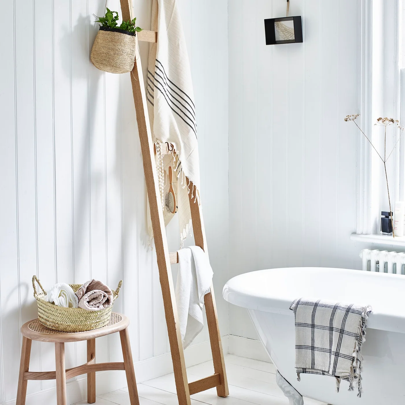 A wooden towel ladder stands beside white paneled walls and a white bath with a small wooden stool and woven basket of towels