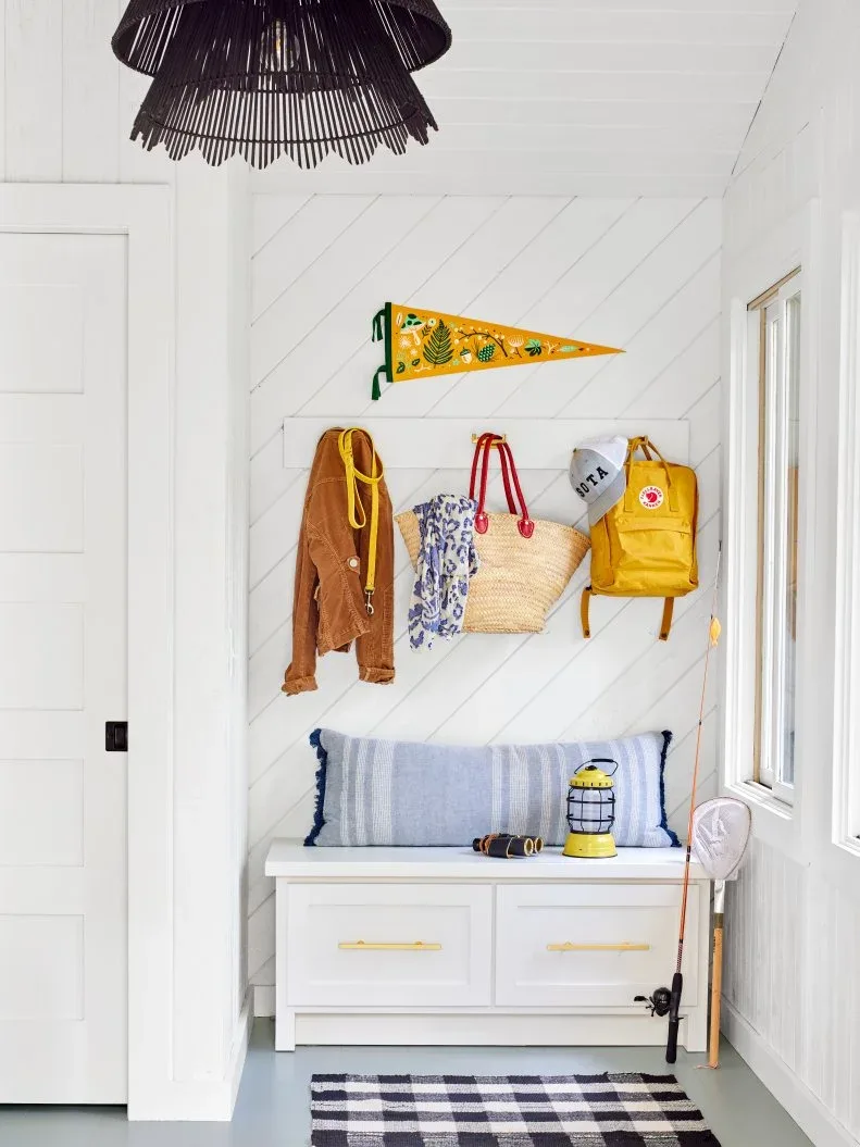 White entry with drawer bench, striped cushion, hanging bags, yellow pennant, dark woven pendant and window.