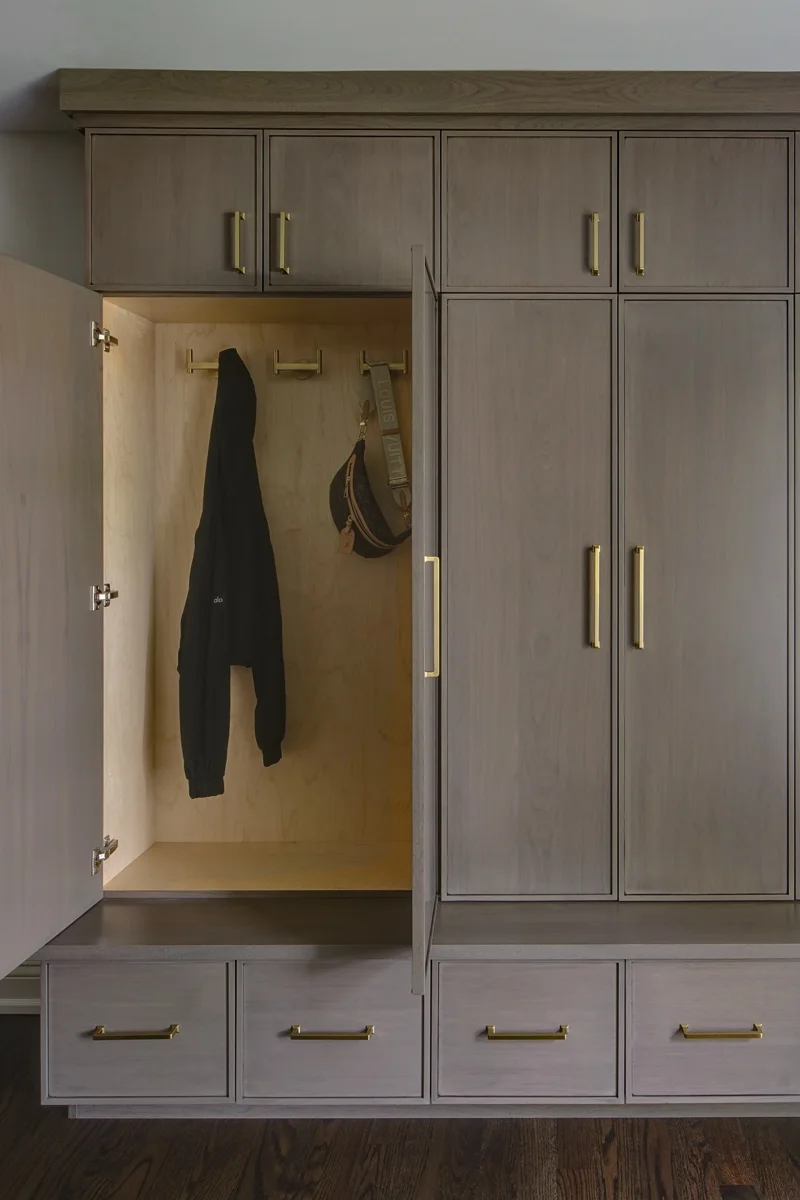 A gray-brown cabinet door opens to a wood-lined coat bay with hooks above drawers, beside matching closed cupboards.