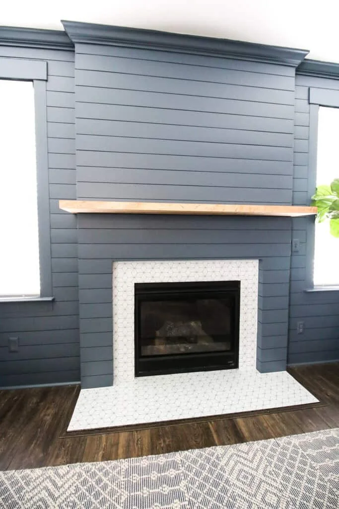 A blue-gray shiplap wall extends around two windows and a projecting fireplace section, with crown molding, a pale wood mantel and a white patterned hearth.
