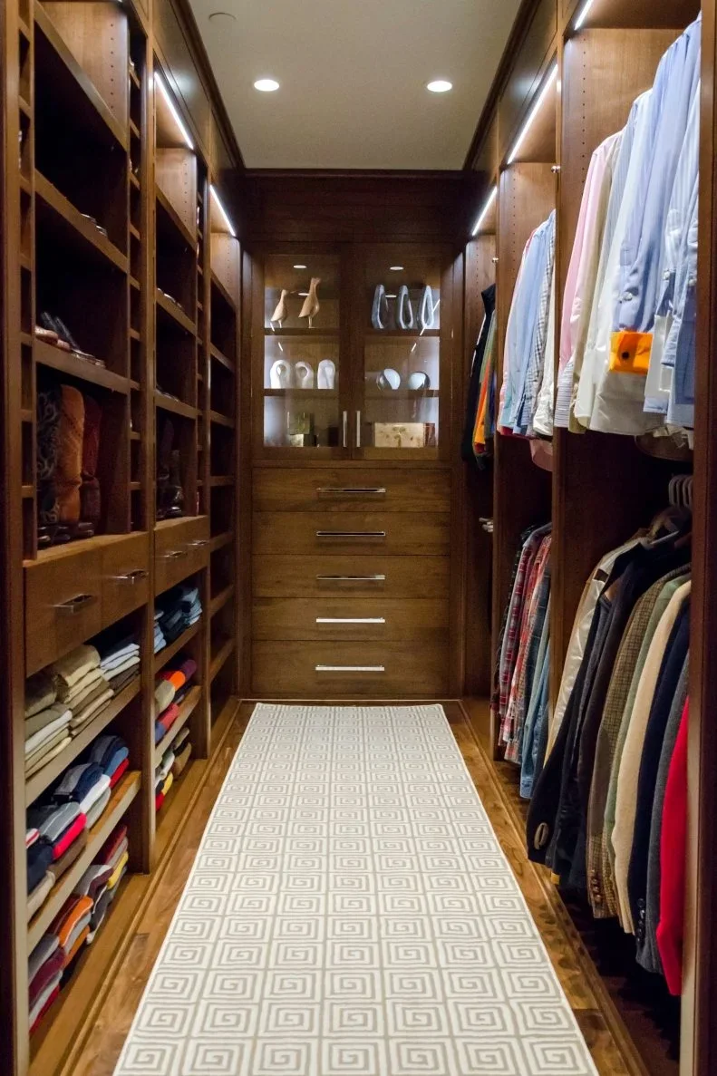 A narrow timber closet has folded clothing and bags on the left, double hanging rails on the right and drawers at the end.