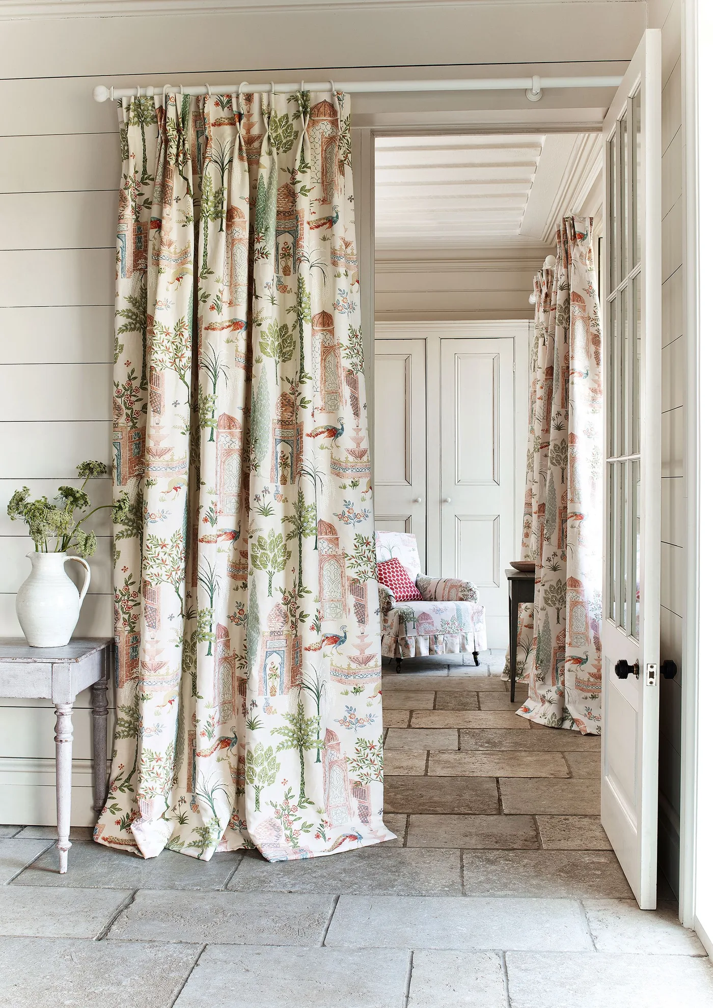 Pale paneled hallway with a full-height patterned curtain, small pale console, jug of flowers and textured stone-like floor.