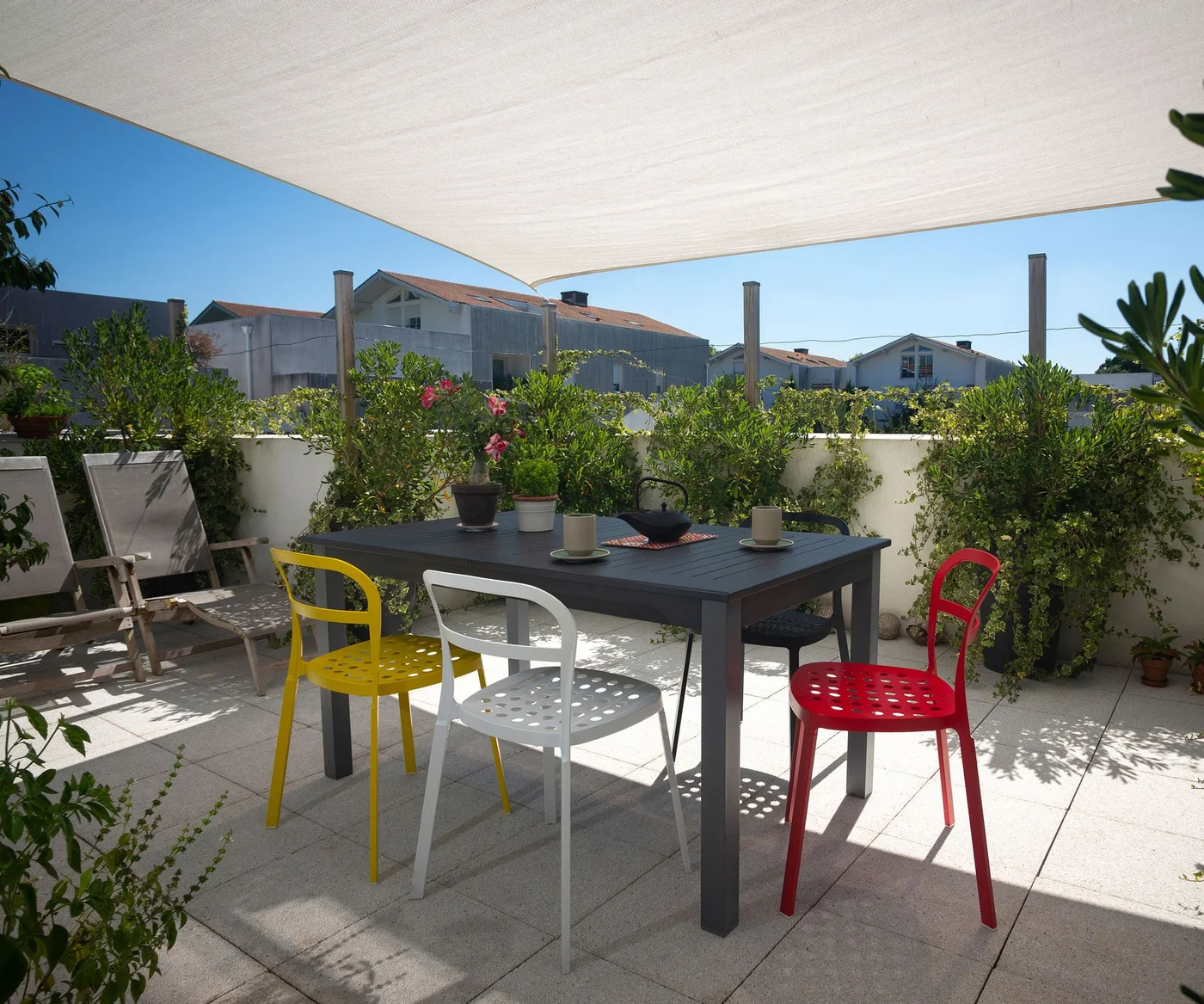 A pale shade sail stretches above a planted terrace dining table with colorful chairs