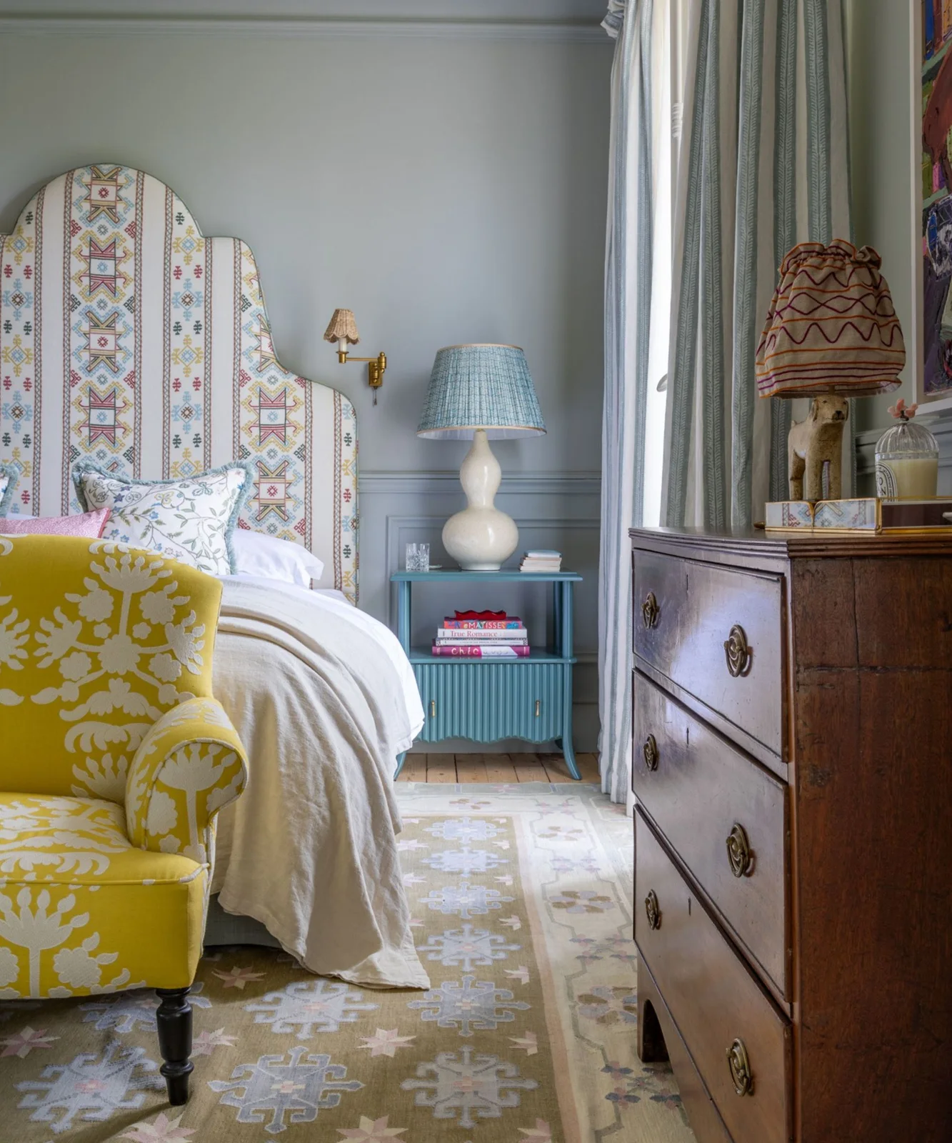 Powder blue bedroom wall and paneling behind a tall patterned headboard with a blue bedside cabinet.