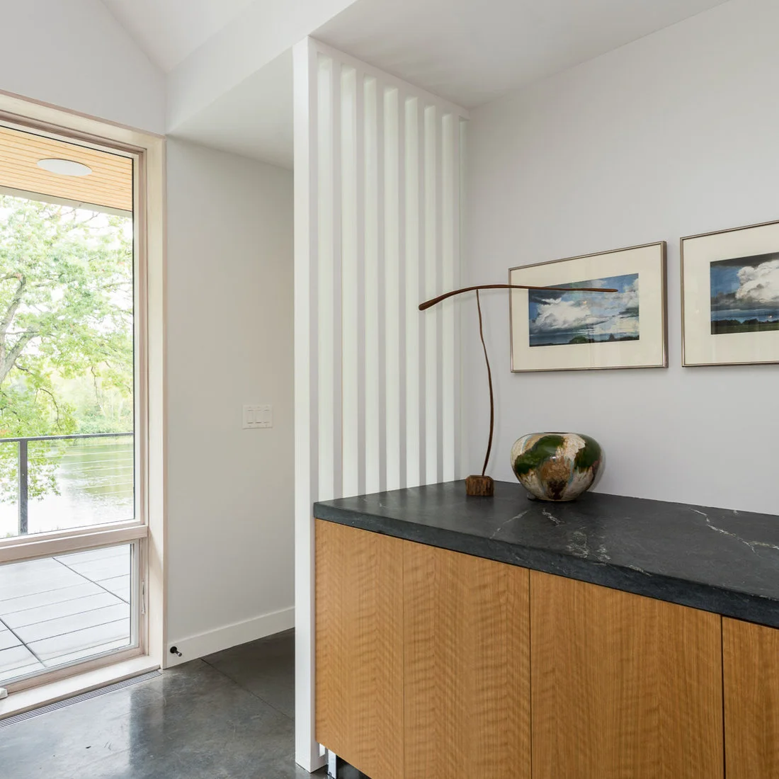 A wood-front cabinet with a dark top stands beneath two landscape pictures beside white vertical wall strips and a tall window.