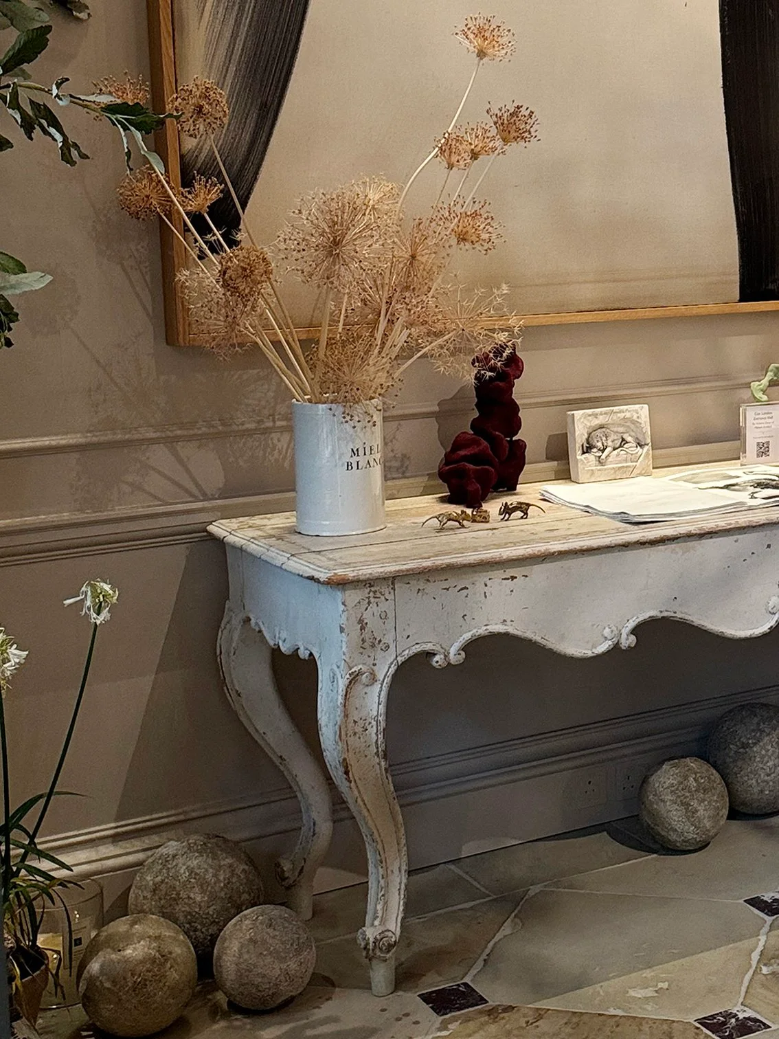 Pale dried seed heads in a light cylindrical tin stand on a distressed pale console beneath a large framed artwork.