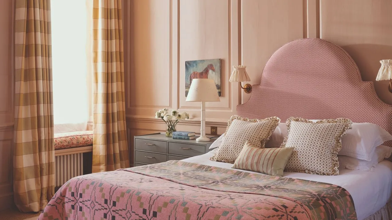 A soft pink curved headboard stands beside modest bedside furniture and patterned bedding.