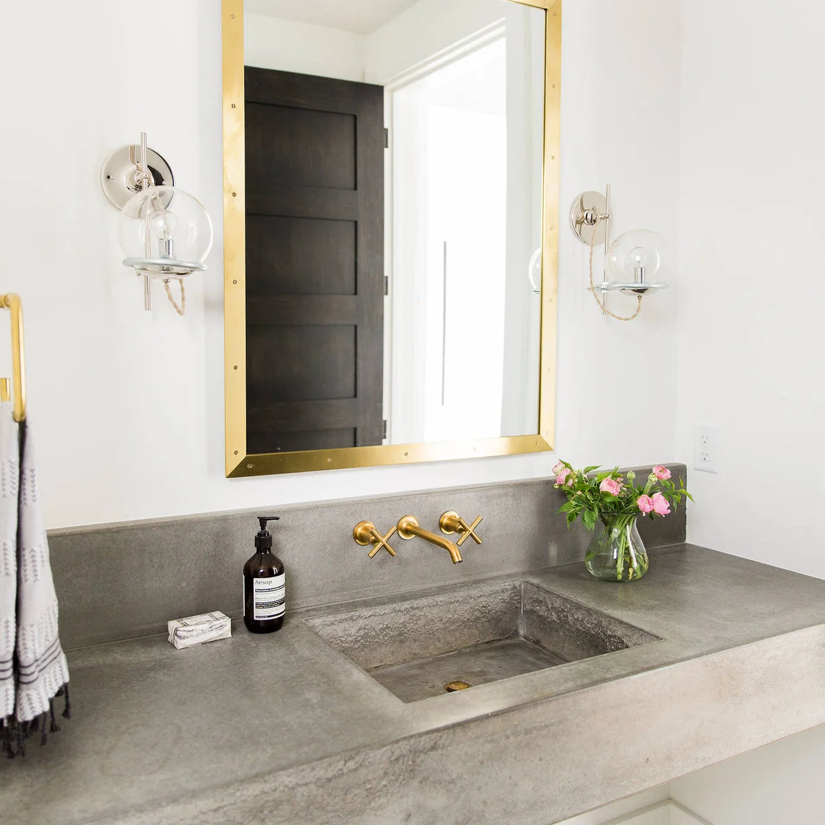 Gray counter with an integrated basin, gold-colored wall taps, amber soap and a clear vase of pink flowers.