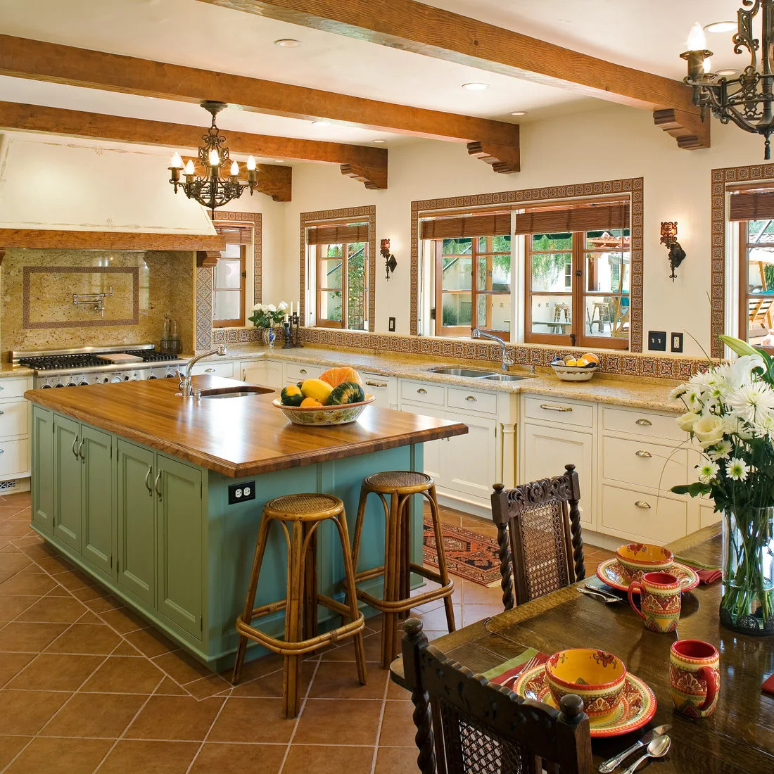 A green island with a timber counter sits beneath beams beside pale cabinets and patterned window borders.