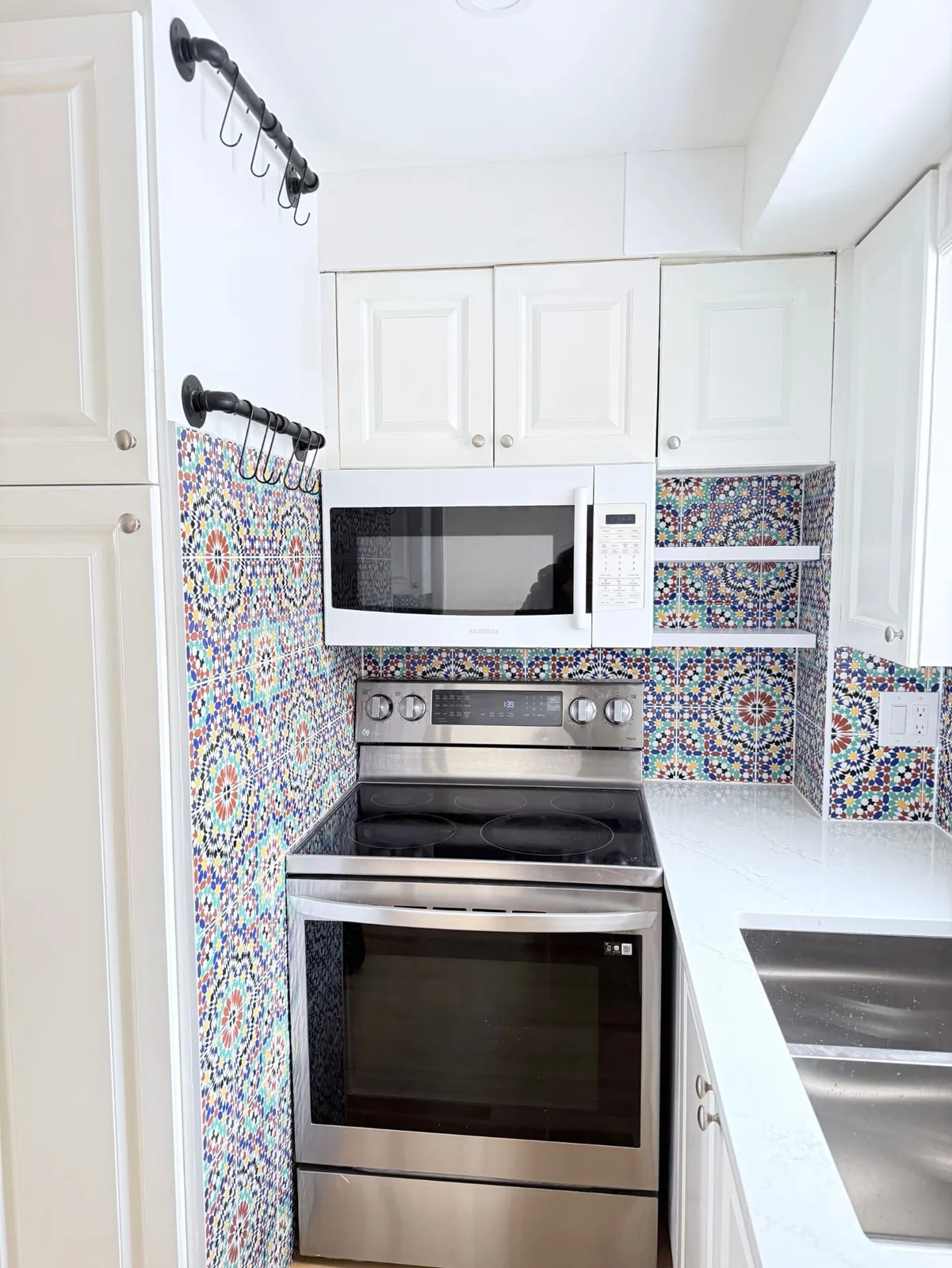 A white galley kitchen has multicolored geometric mosaic patterns around a cooker and microwave beside white cupboards and dark wall-mounted rails