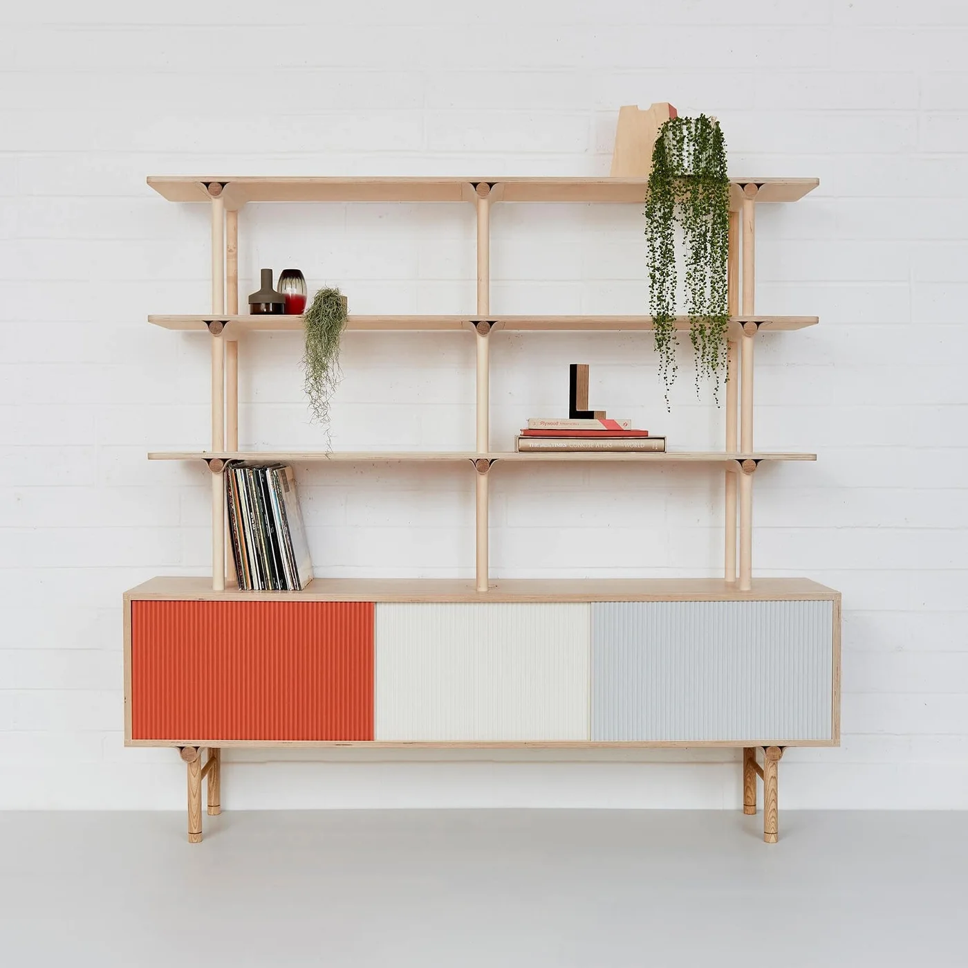 A pale wood sideboard with orange, white and gray ribbed doors supports open shelves with records, two trailing plants, small vessels and books.