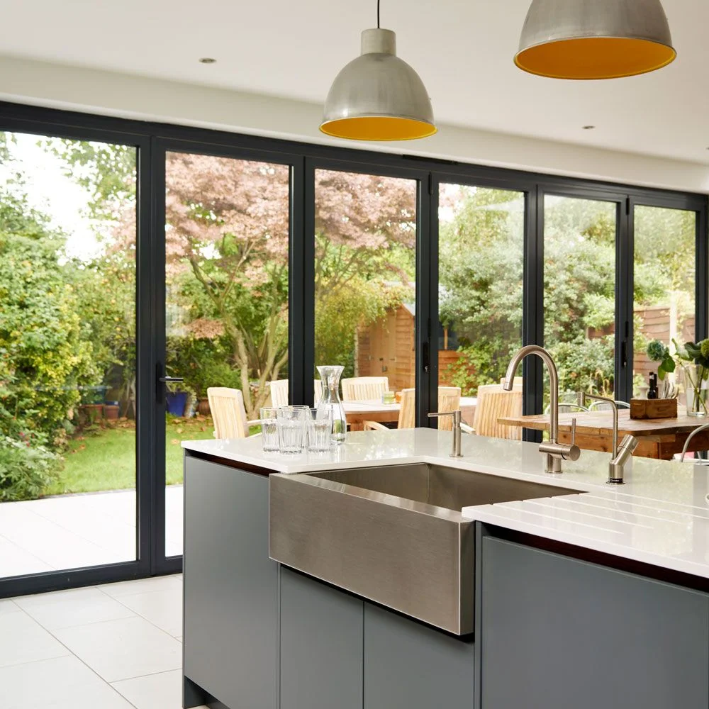 Flat gray cabinet doors sit beneath a broad white counter and deep metal apron-front sink beside full-height garden glazing
