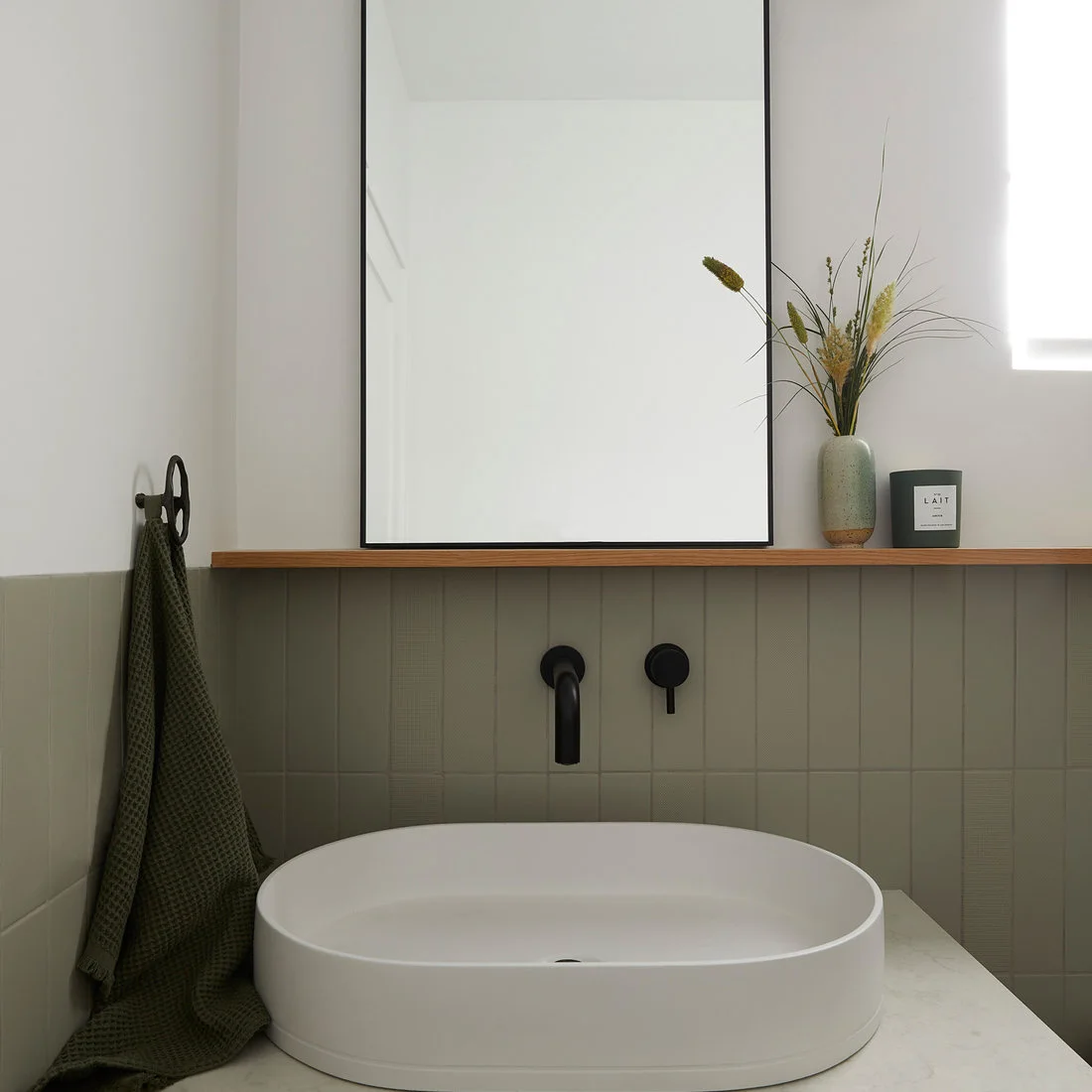 A white oval vessel basin sits below a black wall tap, muted green vertical tiles, thin wood ledge and tall black-edged mirror.